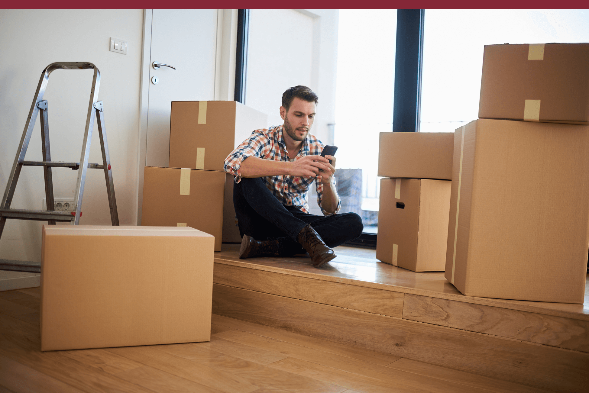 zelle-img-2-optimized.png Man sitting on a wooden floor surrounded by cardboard boxes, looking at his phone during a move. A metal ladder is nearby, and natural light comes in through large windows behind him.